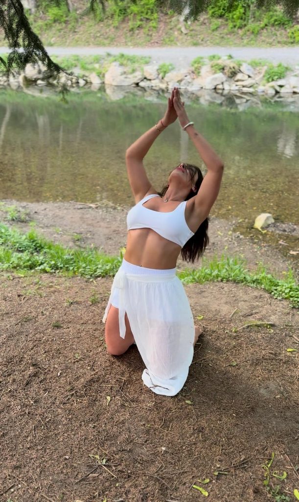 Gera kneeling by the river, arms lifted in prayer pose, dressed in white — embodying connection, serenity, and feminine energy in nature.