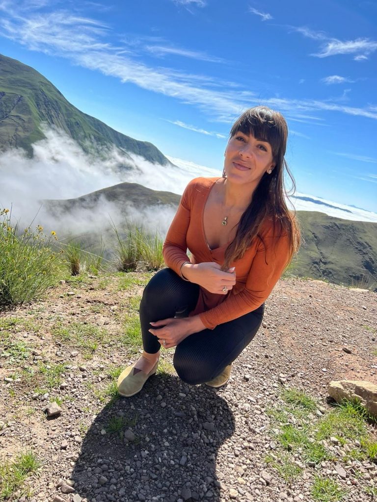 Gera, founder of CoraZONE Fitness, kneeling in nature with mountains and clouds in the background — a moment of reflection and strength.