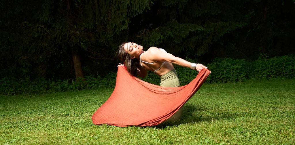 Gera dancing barefoot on the grass, flowing with an orange scarf — channeling grace, strength, and feminine freedom.