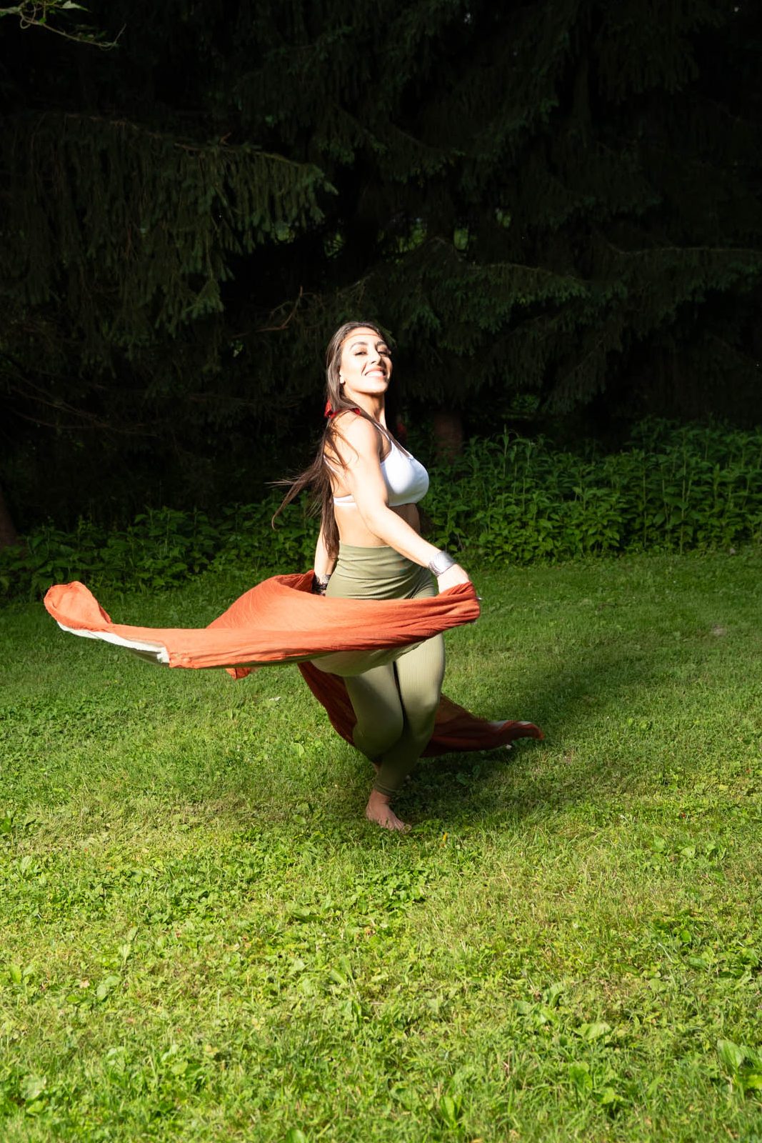 Gera dancing barefoot on the grass, flowing with a burnt orange scarf — expressing joy, freedom, and feminine power.